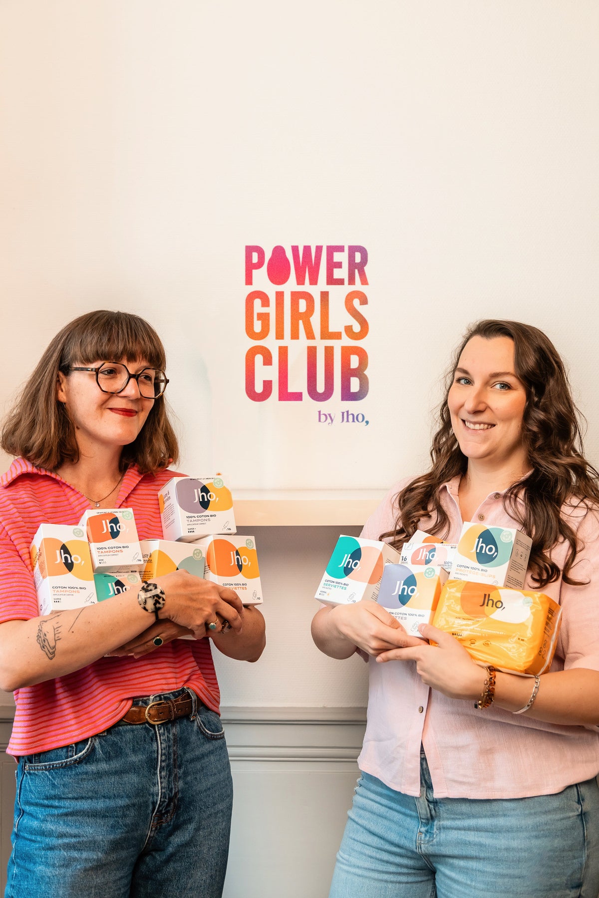 Deux femmes souriantes, les bras chargés de produits Jho, devant le logo Power Girls Club sur le mur