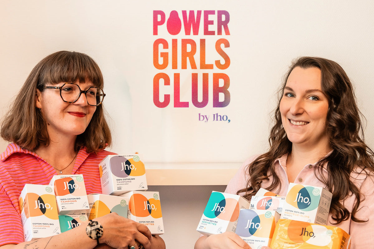Deux femmes souriantes, les bras chargés de produits Jho, devant le logo Power Girls Club